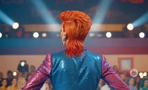 A drag queen with bright orange hair and a sequined blue and pink jacket stands on stage.