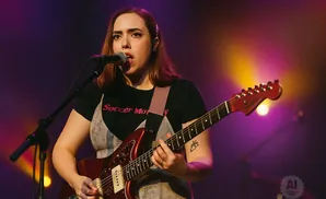 A woman with auburn hair plays a red electric guitar and sings into a microphone on a dimly lit stage.