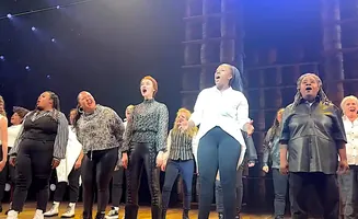 A choir of Black women and girls sing on a dimly lit stage.