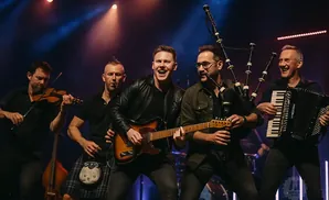 Five men performing music on stage, playing a violin, guitar, bagpipes, and accordion.