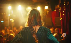 Woman in green velvet dress on stage facing audience, with bright lights and red curtains behind her.
