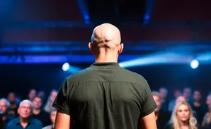 Back of a bald man in a green shirt on stage, facing a blurred audience with blue stage lights.
