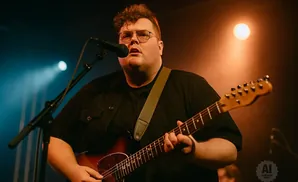 A heavyset man in a black shirt plays a guitar and sings into a microphone on a dimly lit stage.