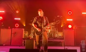 A man plays a black electric guitar on stage, bathed in red and orange stage lights.