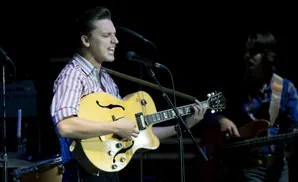 A man in a striped shirt sings and plays a hollow-body electric guitar on stage.