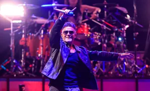 A man in a black leather jacket sings into a microphone on stage, with a drummer in the background.