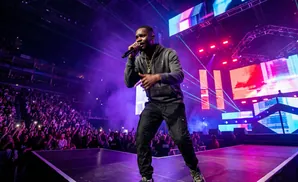 A man in a patterned jacket holds a microphone on a brightly lit stage, with a blurred crowd in the background.