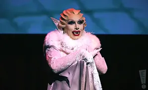 A drag performer with a pale face, sharp fangs, and dramatic makeup wears a fluffy white collar and a light pink outfit with lace gloves.