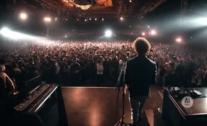 A performer with curly hair faces a cheering crowd from a stage with a keyboard and equipment.