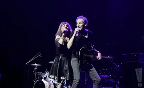 A man and woman sing into microphones on a dark stage with instruments.