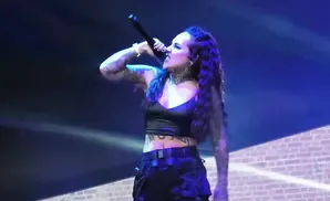 Singer with tattoos and dark curly hair, wearing a black crop top and cargo pants, performs into a microphone on a stage with blue lighting.