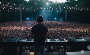DJ with headphones on stage, facing a massive cheering crowd at an outdoor concert.