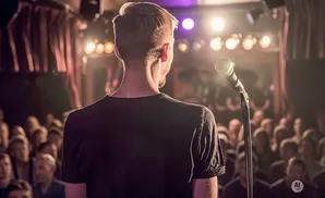 A person stands on stage facing a microphone and an audience in a dimly lit venue.