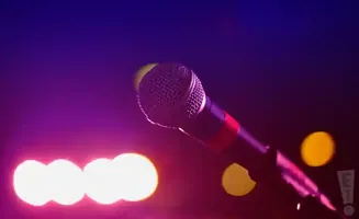 A close-up of a microphone on a stand, illuminated by purple and pink stage lights.