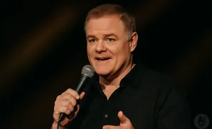 A man in a black shirt holds a microphone and speaks on stage.