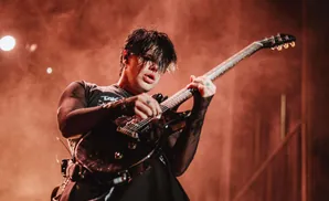 A musician with dark hair plays a black electric guitar on a stage, illuminated by warm, smoky lighting.