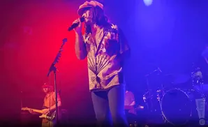 A man in a hat and patterned shirt sings into a microphone on a stage with red and blue lighting.