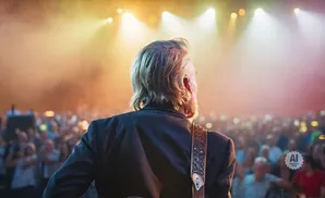 Man with blond hair on stage, back to camera, facing a large, blurred crowd under stage lights.