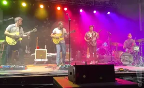 A band performs on a stage with purple and pink lighting. Two guitarists are on the left, and a drummer is on the right.