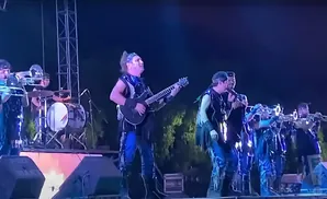 A band performs on stage at night. A guitarist sings while others play brass instruments and drums.