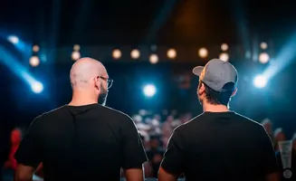 Two men, one bald with glasses and the other wearing a baseball cap, stand with their backs to the camera facing a stage with bright lights.