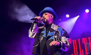 Boy George sings into a microphone on stage, wearing a blue hat and a colorful jacket under purple lights.