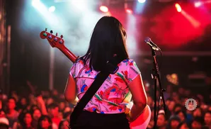 Woman with red guitar on stage in front of a cheering crowd, bathed in colorful stage lights.