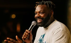 A Black man with dreadlocks and a beard laughs while holding a microphone and gesturing with his hand.