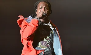 A rapper with dreadlocks, wearing a red jacket and a chain, performs into a microphone.