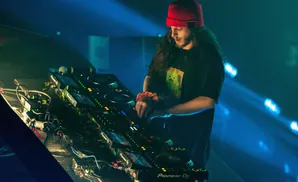 DJ with long dark hair and red beanie, performing on Pioneer DJ equipment under blue stage lights.
