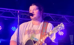 A man with a mustache sings into a microphone while playing a guitar on stage under blue and purple lights.