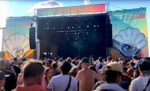 A crowded outdoor concert stage with banners, lights, and a large shell graphic with an eye.