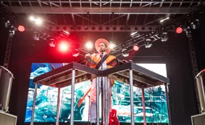 Musician in a hat and orange jacket plays a keyboard on stage with a large screen behind him.
