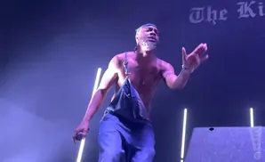A shirtless man with overalls hanging down performs on stage under purple lights.