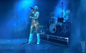 A woman in a gold outfit and white boots sings into a microphone on a stage with a drum set behind her.