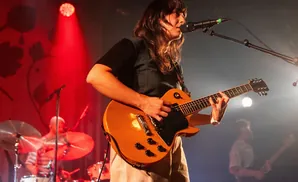 A woman plays an orange electric guitar while singing into a microphone on a dimly lit stage.
