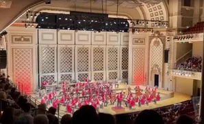 An orchestra performs in a grand concert hall with intricate white wall paneling and an audience in the foreground.