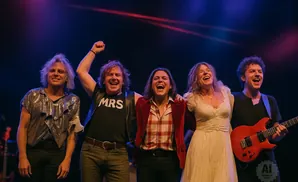 Five band members laugh on stage, with two playing guitar.