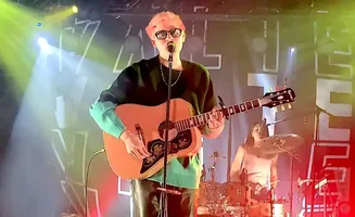 A musician with pink hair plays an acoustic guitar on stage, with a drummer in the background.