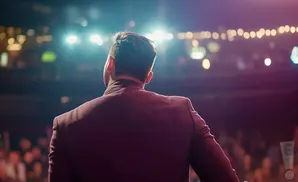 Man in a maroon suit facing away from the camera on a stage, with bright stage lights and an audience in the background.
