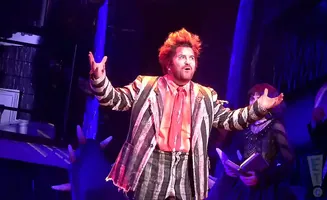 A man with spiky orange hair in a striped suit and tie, on stage with other performers.