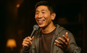 A man laughs into a microphone while gesturing with his hands, wearing a gray hoodie over a brown shirt.