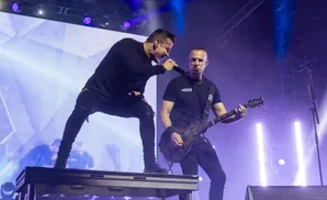 A male singer and guitarist perform on a brightly lit stage with purple lights and a large screen.