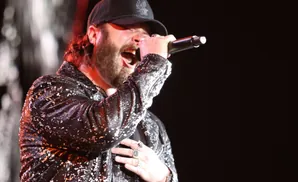 A man with a beard sings into a microphone, wearing a sparkly jacket and a black baseball cap.