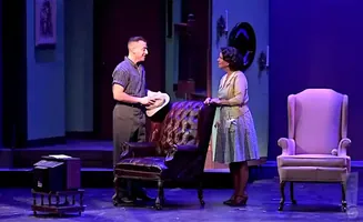 Two people stand on a stage. A man in a striped shirt and gray pants holds a hat, and a woman in a cardigan and checkered dress stands beside him.