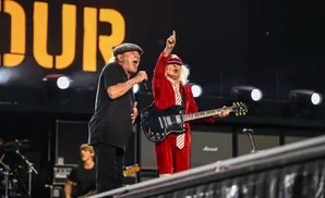 Two male musicians perform on stage, one singing into a microphone, the other playing a guitar.