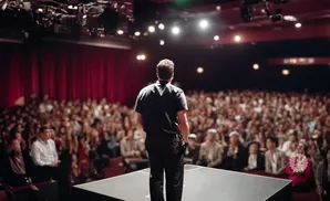 A man in a black shirt stands on a stage facing a large audience in a theater.