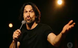 A man with dark hair and a beard holds a microphone and gestures with his right hand.