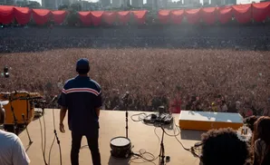 A man in a blue shirt and hat stands on a stage facing a massive, cheering crowd.