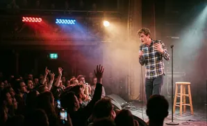 A comedian performs on stage with a microphone, while the audience cheers with hands raised.